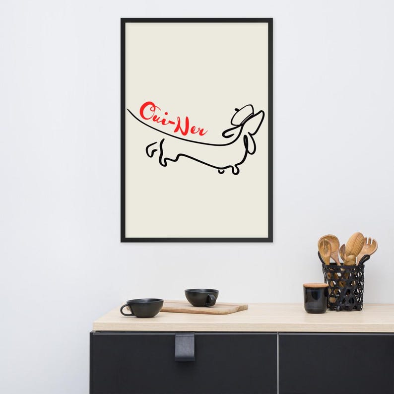 May include: Framed art print featuring a minimalist black line drawing of a dachshund wearing a hat, with the word "Cui-Ner" in red script. The artwork is set against a cream background, enclosed in a black frame.