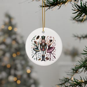 Könnte beinhalten: Ein weißer, runder Weihnachtsschmuck mit einem Nussknacker und zwei Ballerinas. Der Nussknacker trägt eine grüne und rote Uniform. Die Ballerinas tragen Tutus in Lila und Türkis. Der Schmuck hängt an einem goldenen Faden.
