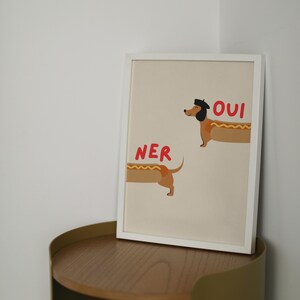 K&ouml;nnte beinhalten: Ein gerahmter Kunstdruck mit einer skurrilen Illustration von zwei Dackeln, die als Hotdogs gestaltet sind. Einer der Hunde tr&auml;gt einen Hut, mit dem Wort "OUI" in Rot. Das Wort "NER" steht in Rot neben dem anderen Hund.