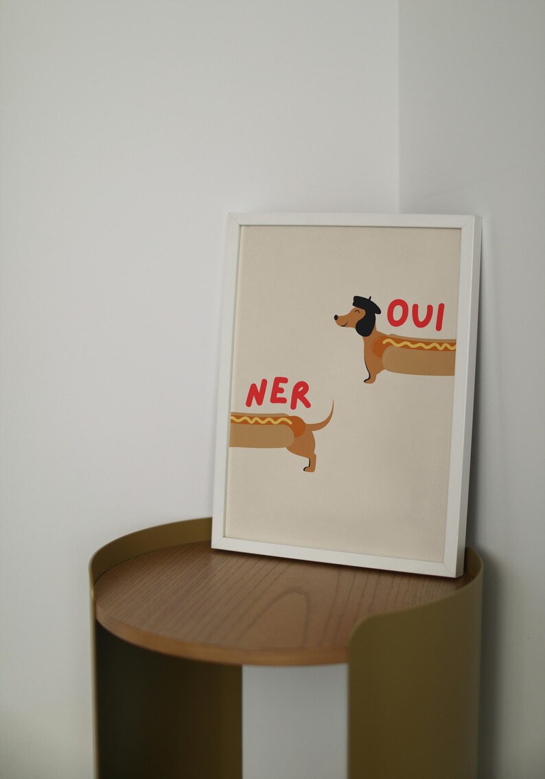 K&ouml;nnte beinhalten: Ein gerahmter Kunstdruck mit einer skurrilen Illustration von zwei Dackeln, die als Hotdogs gestaltet sind. Einer der Hunde tr&auml;gt einen Hut, mit dem Wort "OUI" in Rot. Das Wort "NER" steht in Rot neben dem anderen Hund.