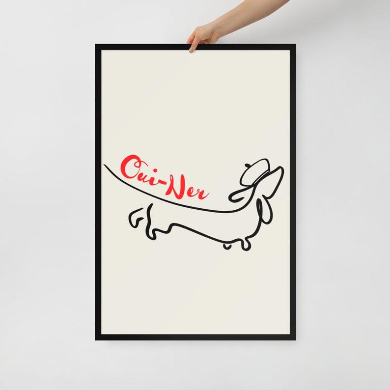 May include: Framed art print featuring a minimalist line drawing of a dachshund dog in black ink. The word "Oui-Ner" is written in red cursive above the dog. The background is a cream color, and the frame is black.