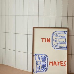 Pu&ograve; includere: Una stampa incorniciata con sfondo crema e cornice in legno marrone. La stampa presenta testo blu e rosso che recita "TIN MATES" e "SARDINE". L'immagine include un'illustrazione stilizzata di sardine in scatola.