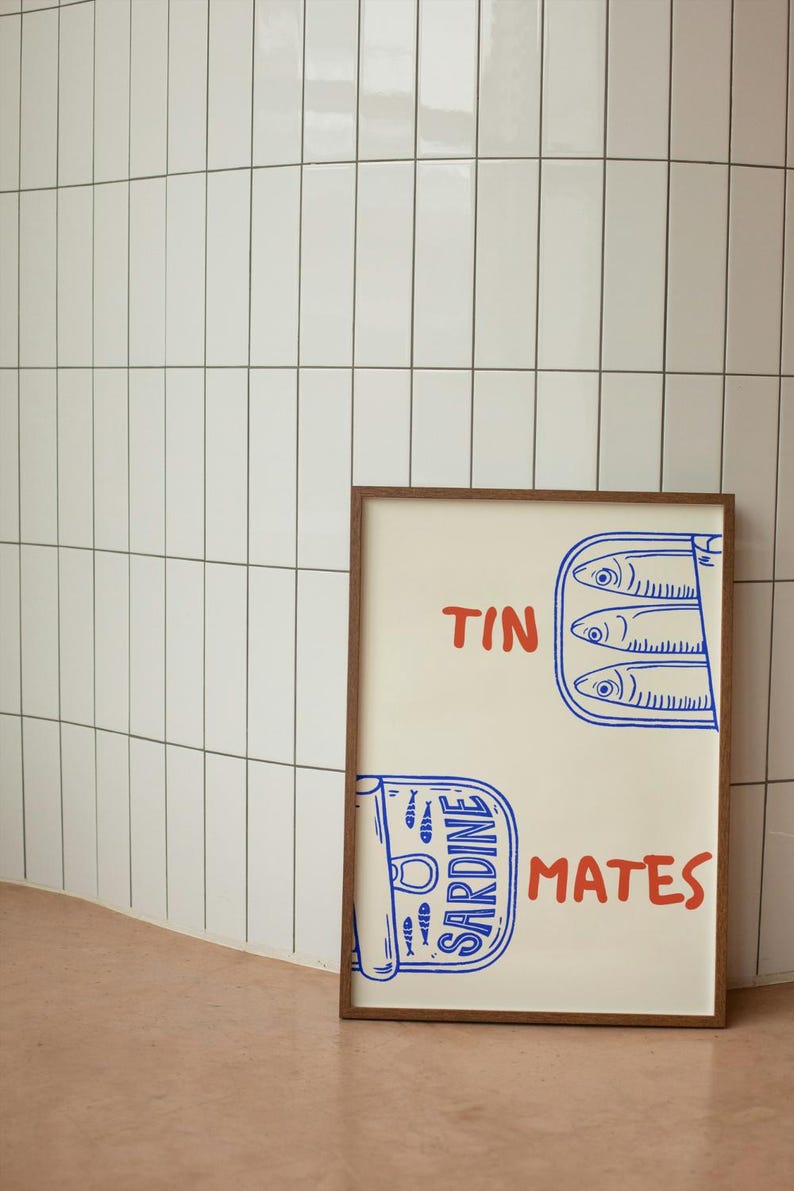 Pu&ograve; includere: Una stampa incorniciata con sfondo crema e cornice in legno marrone. La stampa presenta testo blu e rosso che recita "TIN MATES" e "SARDINE". L'immagine include un'illustrazione stilizzata di sardine in scatola.