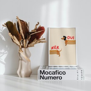K&ouml;nnte beinhalten: Ein gerahmter Kunstdruck mit zwei Dackeln in Hotdog-Br&ouml;tchen, den Worten "Oui" und "Ner" in Rot. Eine beige Vase h&auml;lt getrocknete Blumen. Der Druck liegt auf einem Stapel B&uuml;cher mit dem Titel "Mocafico Numero".