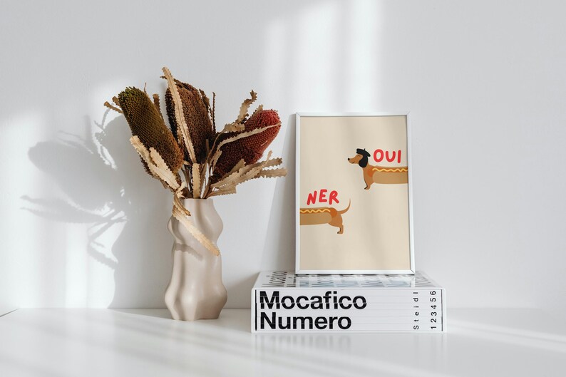 K&ouml;nnte beinhalten: Ein gerahmter Kunstdruck mit zwei Dackeln in Hotdog-Br&ouml;tchen, den Worten "Oui" und "Ner" in Rot. Eine beige Vase h&auml;lt getrocknete Blumen. Der Druck liegt auf einem Stapel B&uuml;cher mit dem Titel "Mocafico Numero".