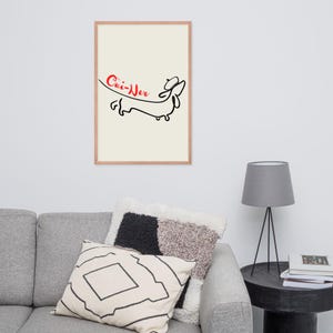 May include: A framed art print featuring a minimalist line drawing of a dachshund dog wearing a beret. The dog is black, and the text "Cui-Ner" is written in red cursive above it. The background is off-white, and the frame is light brown.