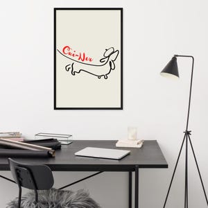 May include: Black framed art print featuring a minimalist line drawing of a dachshund wearing a hat. The red text above the dog reads "Cui-Wer" on a cream background. The artwork is displayed on a black desk.
