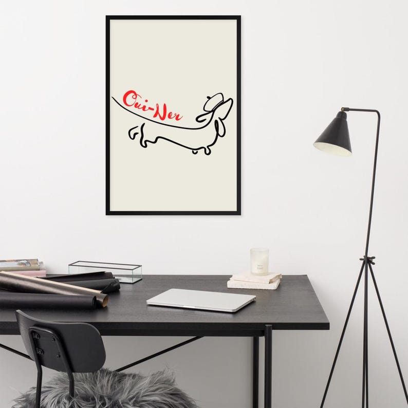 May include: Black framed art print featuring a minimalist line drawing of a dachshund wearing a hat. The red text above the dog reads "Cui-Wer" on a cream background. The artwork is displayed on a black desk.