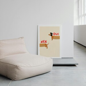 K&ouml;nnte beinhalten: Ein beiger Sitzsack steht neben einem gerahmten Druck. Der Druck zeigt zwei Cartoon-Dackel in Hotdog-Br&ouml;tchen mit den roten W&ouml;rtern "NER" und "OUI". Der Hintergrund ist hellbeige.