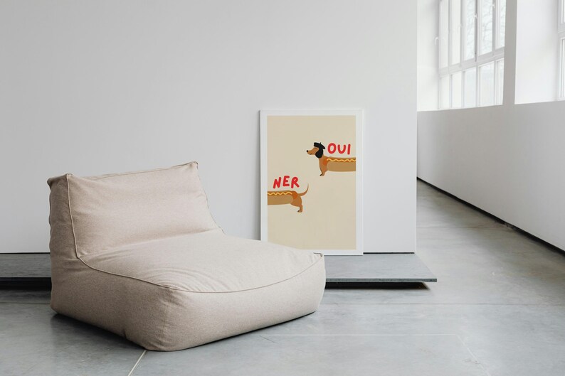 K&ouml;nnte beinhalten: Ein beiger Sitzsack steht neben einem gerahmten Druck. Der Druck zeigt zwei Cartoon-Dackel in Hotdog-Br&ouml;tchen mit den roten W&ouml;rtern "NER" und "OUI". Der Hintergrund ist hellbeige.