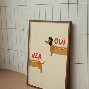K&ouml;nnte beinhalten: Ein gerahmter Kunstdruck mit einer Cartoon-Illustration von zwei Dackeln als Hotdogs. Einer der Hunde tr&auml;gt ein Barett und das Wort "OUI" steht dar&uuml;ber. Der andere Hund hat das Wort "NER" dar&uuml;ber. Der Hintergrund ist hellbeige.