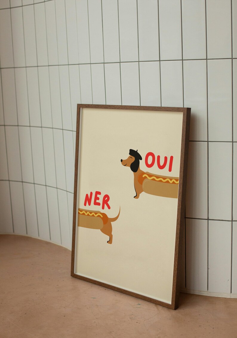 K&ouml;nnte beinhalten: Ein gerahmter Kunstdruck mit einer Cartoon-Illustration von zwei Dackeln als Hotdogs. Einer der Hunde tr&auml;gt ein Barett und das Wort "OUI" steht dar&uuml;ber. Der andere Hund hat das Wort "NER" dar&uuml;ber. Der Hintergrund ist hellbeige.
