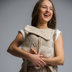 May include: A woman wearing a white crocheted top and beige trousers. She is holding a beige crocheted bag in front of her.