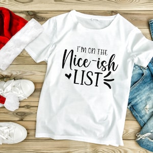 May include: White t-shirt with black text that reads "I'm on the Nice-ish List" with a heart and a squiggly line. The shirt is laid out on a wooden surface with a red Santa hat, white trainers, and blue jeans.