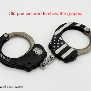 May include: A pair of black handcuffs with silver-colored metal rings and a connecting chain. One cuff features a white and black American flag design, while the other has the name "Freeman" in white lettering. The text "Old pair pictured to show the graphic" is visible.