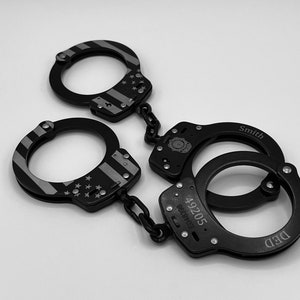 May include: Black handcuffs with a silver and black American flag design on the cuffs. One pair has the name "Smith" engraved, and another pair has the numbers "49205" and "ED48398" engraved. The handcuffs are connected by a chain.