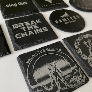Op de afbeelding: Een verzameling zwarte leistenen onderzetters met verschillende geëtste ontwerpen en tekst. Sommige onderzetters tonen zinnen als "Break the Chains" en "Howlers". De onderzetters hebben een natuurlijke, ruwe rand en een donkergrijze kleur.