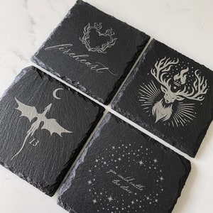 May include: Set of four black slate coasters with white engraved designs. The designs include a heart with flames, a stag with antlers, a dragon, and a constellation with the text "you could rattle the stars".