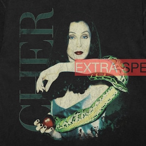 Może przedstawiać: Czarno-biały vintage'owy t-shirt z portretem Cher z wężem owiniętym wokół jej ramienia. Tekst "CHER" jest nadrukowany pionowo po lewej stronie obrazu. Tekst "EXTRA SPE" jest częściowo widoczny w prawym górnym rogu obrazu.