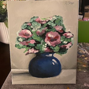 May include: A painting of a bouquet of pink flowers in a blue vase. The flowers are painted in a loose, impressionistic style. The vase is a simple, round shape. The painting is signed "P. Skaggs" in the upper right corner.