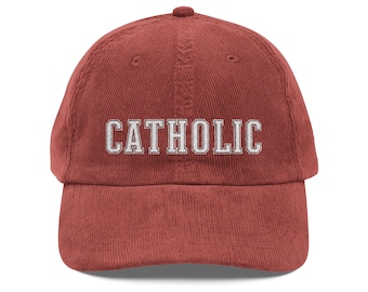May include: A brick-red corduroy baseball cap with the word "CATHOLIC" embroidered in white block letters across the front. The cap has a curved brim and an adjustable strap.