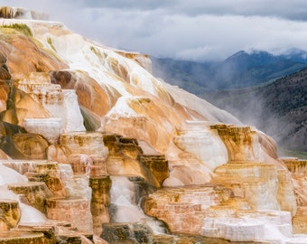 May include: A scenic landscape of a terraced, mineral-rich hillside with cascading water. The formations display shades of white, beige, and brown. The background features mountains and a cloudy sky, creating a natural, serene environment.