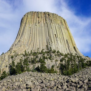May include: A large, towering rock formation with a light brown color and a striated texture. The rock is surrounded by a rocky landscape and some trees.