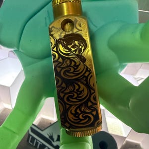 May include: A gold-colored, rectangular lighter with intricate black and gold swirling patterns and a detailed illustration. The lighter has a textured top and bottom, and a small circular hole. The lighter is held by a green object.