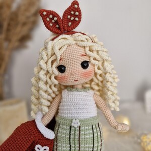 Op de afbeelding: Een gehaakte pop met blond krullend haar, die een wit tanktopje, een groene geruite rok en een rood hoofdband met een bruin bladerenontwerp draagt.