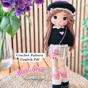 Może przedstawiać: Ręcznie robiona szydełkowa lalka z długimi brązowymi włosami, w czarnym berecie, swetrze i plisowanej spódnicy. Lalka ma różową torebkę w kształcie serca oraz różowe i czarne buty. Widoczny jest tekst "Crochet Pattern English Pdf" i "Meet Aria".