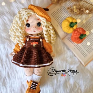 Pode incluir: Uma boneca de croch&ecirc; com cabelo loiro encaracolado usando um vestido marrom, um bon&eacute; marrom e botas laranja. A boneca est&aacute; sentada em uma superf&iacute;cie branca e felpuda com um livro e duas pequenas ab&oacute;boras laranja no fundo. O texto "_Organic_Toyss_" &eacute; vis&iacute;vel no canto inferior direito da imagem.