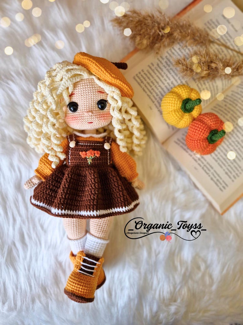 Pode incluir: Uma boneca de croch&ecirc; com cabelo loiro encaracolado usando um vestido marrom, um bon&eacute; marrom e botas laranja. A boneca est&aacute; sentada em uma superf&iacute;cie branca e felpuda com um livro e duas pequenas ab&oacute;boras laranja no fundo. O texto "_Organic_Toyss_" &eacute; vis&iacute;vel no canto inferior direito da imagem.