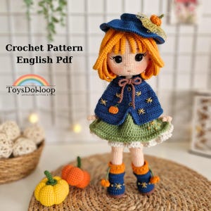 May include: A handmade crochet doll with orange hair, wearing a blue cape with star and pumpkin details, a green dress, and blue boots. The doll is part of a crochet pattern, as indicated by the text "Crochet Pattern English Pdf".