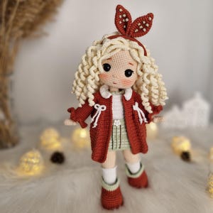 Pode incluir: Uma boneca de croch&ecirc; com cabelo loiro encaracolado vestindo um casaco vermelho, uma saia verde e meias brancas. A boneca tem uma faixa de cabe&ccedil;a branca com bolinhas vermelhas e olhos castanhos.