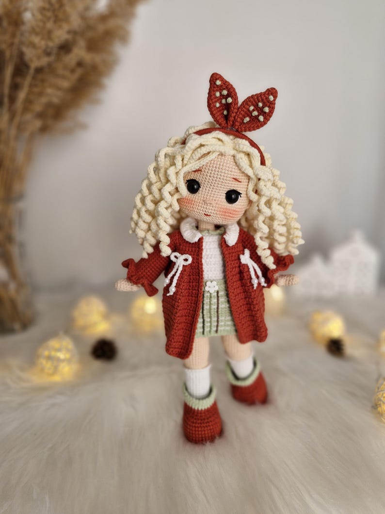 Pode incluir: Uma boneca de croch&ecirc; com cabelo loiro encaracolado vestindo um casaco vermelho, uma saia verde e meias brancas. A boneca tem uma faixa de cabe&ccedil;a branca com bolinhas vermelhas e olhos castanhos.