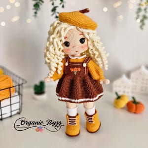 Pode incluir: Uma boneca de croch&ecirc; com cabelo loiro encaracolado vestindo um vestido marrom com uma borda branca, um bon&eacute; amarelo e botas amarelas. A boneca est&aacute; em p&eacute; sobre uma superf&iacute;cie branca com uma pequena planta em vaso e duas ab&oacute;boras no fundo. O texto "_Organic_Toyss_" &eacute; vis&iacute;vel no canto inferior esquerdo da imagem.