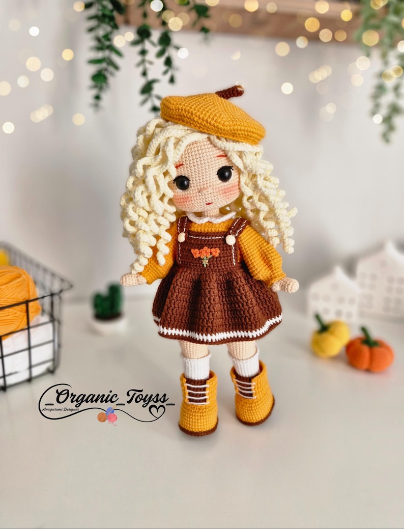 Pode incluir: Uma boneca de croch&ecirc; com cabelo loiro encaracolado vestindo um vestido marrom com uma borda branca, um bon&eacute; amarelo e botas amarelas. A boneca est&aacute; em p&eacute; sobre uma superf&iacute;cie branca com uma pequena planta em vaso e duas ab&oacute;boras no fundo. O texto "_Organic_Toyss_" &eacute; vis&iacute;vel no canto inferior esquerdo da imagem.