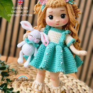May include: A crocheted doll with blonde hair, wearing a teal dress and holding a white bunny. The doll has a sweet expression and is adorned with pink flower accents. The text "ELORA & NIA ENGLISH PDF CROCHET PATTERN" is visible.