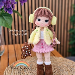 Puede incluir: Muñeca de ganchillo hecha a mano con cabello castaño, que lleva una chaqueta amarilla, una falda rosa y botas marrones. La muñeca tiene una pequeña mochila y auriculares. El texto "crochet pattern english pdf" es visible.