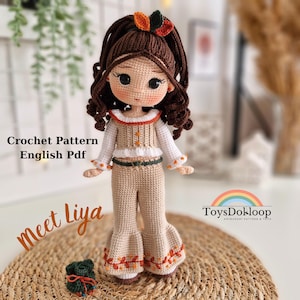 以下が含まれることがあります： 手作りの編み人形、Liya。茶色の髪とボヘミアンスタイルの衣装を着ています。人形はクリーム色のトップスとオレンジ色のアクセントが付いたパンツを着用しています。「Crochet Pattern English Pdf」と「Meet Liya」の文字が見えます。