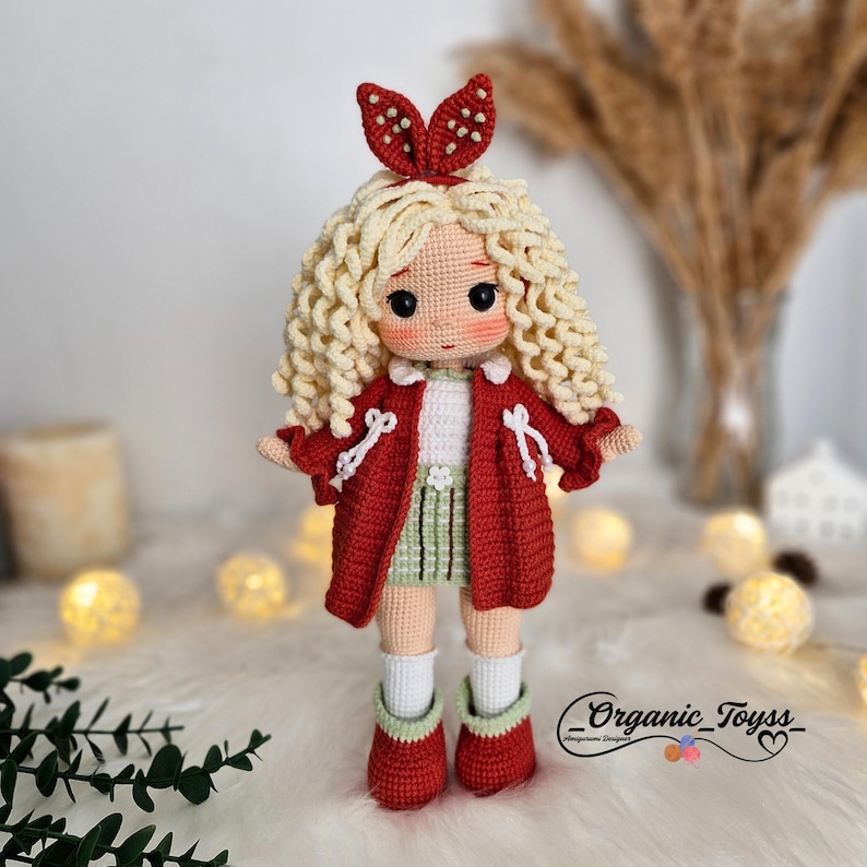 Pode incluir: Uma boneca de croch&ecirc; com cabelo loiro encaracolado usando uma jaqueta vermelha, uma saia verde e sapatos vermelhos. A boneca tem um la&ccedil;o vermelho na cabe&ccedil;a.