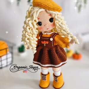 May include: A crocheted doll with blonde curly hair, wearing a brown dress with a white trim, a yellow beret, and yellow boots. The doll is standing on a white surface.  _Organic_Toyss_ is written on the bottom of the image.