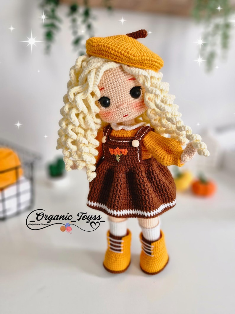 May include: A crocheted doll with blonde curly hair, wearing a brown dress with a white trim, a yellow beret, and yellow boots. The doll is standing on a white surface.  _Organic_Toyss_ is written on the bottom of the image.