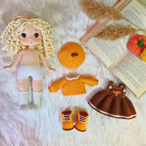 May include: A crocheted doll with blonde curly hair wearing a white dress and white boots. The doll is surrounded by crocheted clothing items, including a brown dress, a yellow sweater, a yellow hat, and a pair of yellow boots.