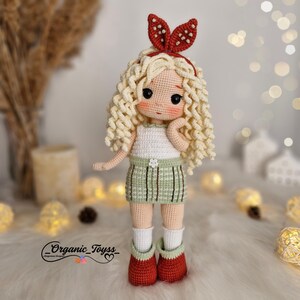 Op de afbeelding: Een gehaakte pop met blond krullend haar, die een groen-wit geruite rok, een wit tanktopje en een rood hoofdband met witte stippen draagt. De pop draagt rode schoenen met witte sokken.