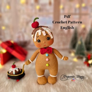 Puede incluir: Patrón de crochet para una muñeca de jengibre con un lazo rojo, glaseado blanco y una cereza en la parte superior. La muñeca sostiene un pequeño postre de jengibre con una cereza en la parte superior. El patrón está en inglés y está disponible para descargar en PDF.