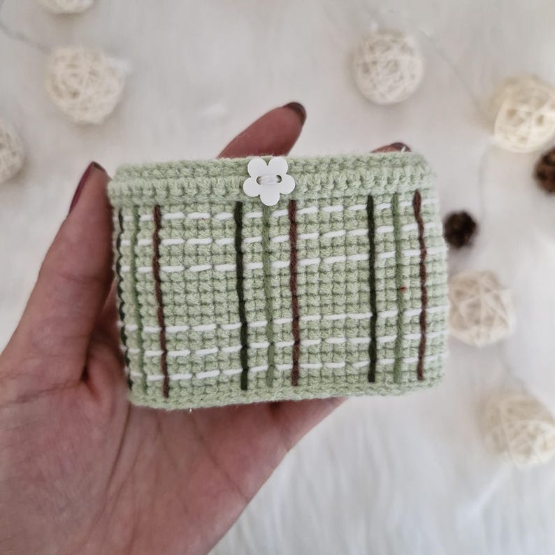 Op de afbeelding: Een klein groen gehaakt mandje met witte en bruine strepen. Het mandje heeft een witte, bloemvormige knoop bovenop.