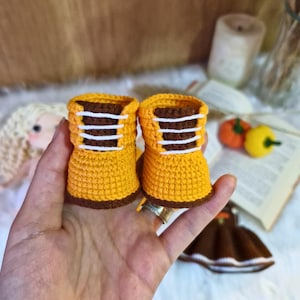 May include: A pair of yellow crocheted booties with brown and white trim. The booties have a lace-up design.