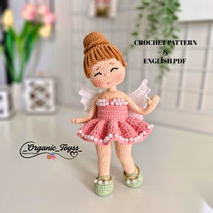 May include: Crochet pattern for a pink ballerina doll with white wings and a pearl necklace. The doll has brown hair and a sweet expression. The pattern is available in English PDF format.