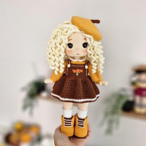 May include: A crocheted doll with blonde curly hair, wearing a brown dress with a white trim, a yellow beret, and yellow boots.